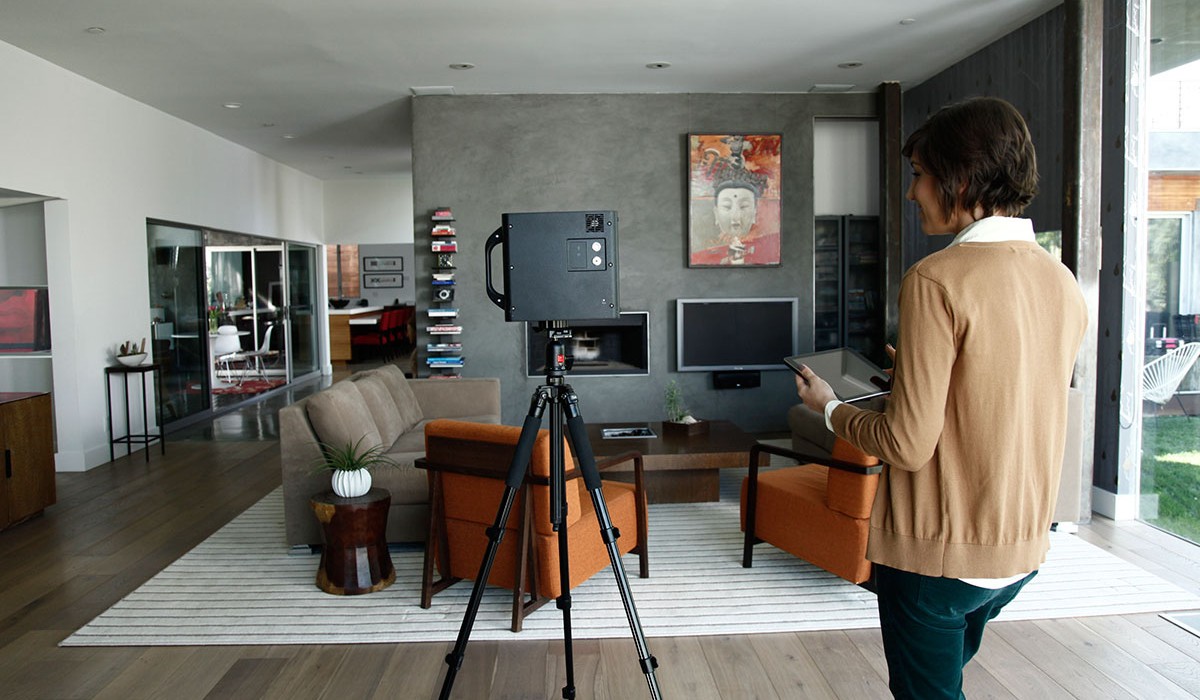 A 3D tour camera on a tripod scanning a living and dining area while an agent controls it from a tablet
