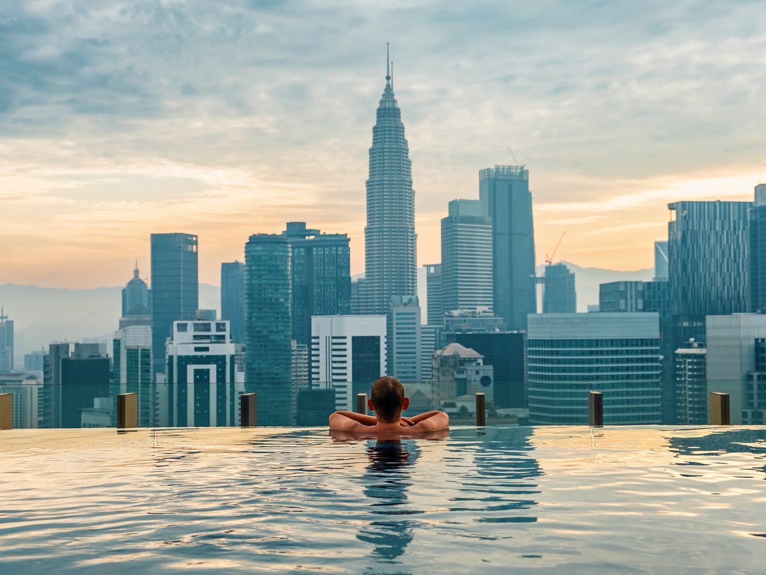 A visitor resting at the edge of a rooftop infinity pool at sunrise, looking out over the Kuala Lumpur skyline and the Petronas Twin Towers